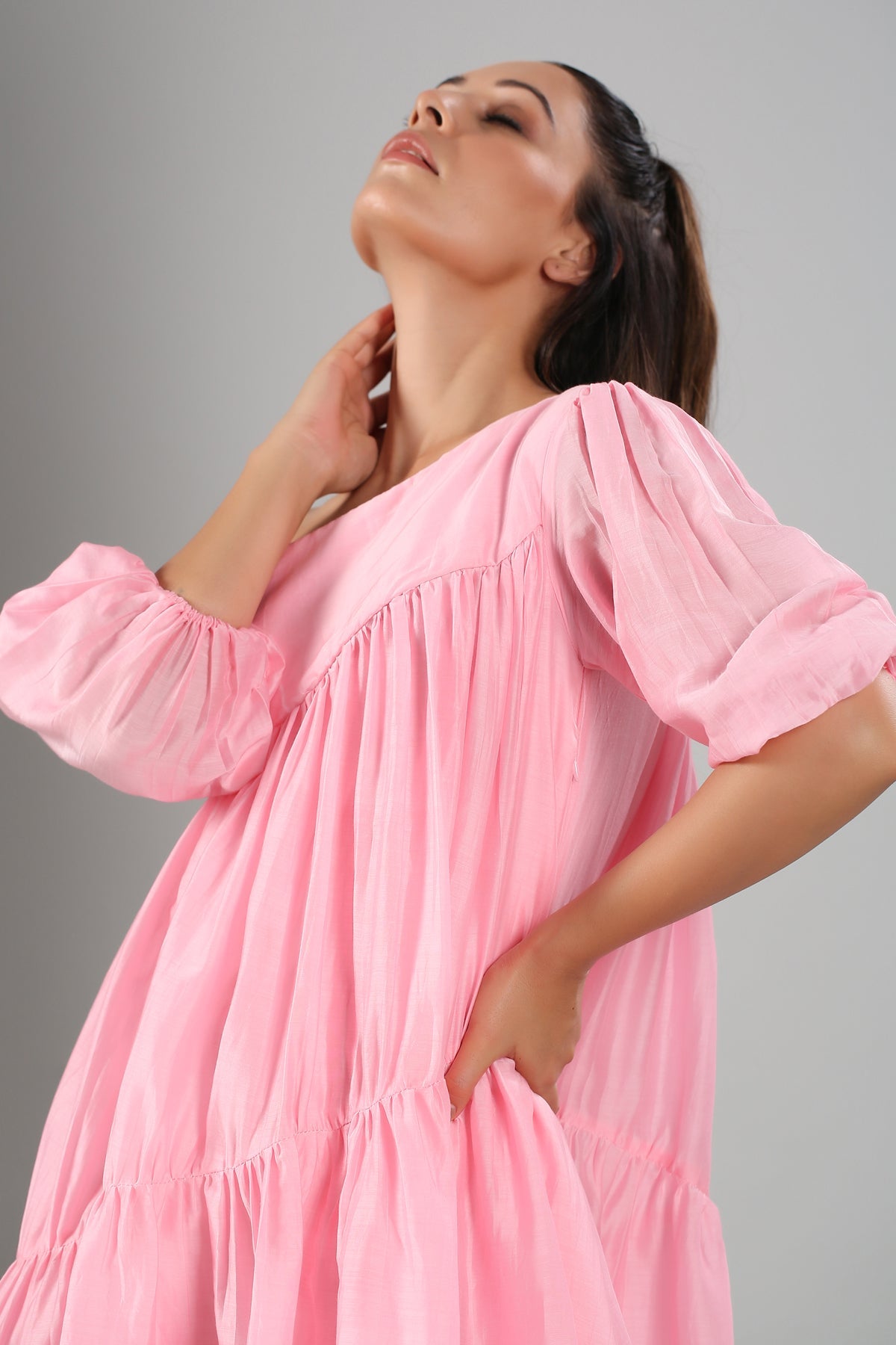 Model in Stylera pink off shoulder dress with puff sleeves and tiered skirt posing with hand at neck