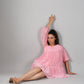 Woman seated against gray backdrop wearing a pink off shoulder dress in flowy tiered mini style and sandals