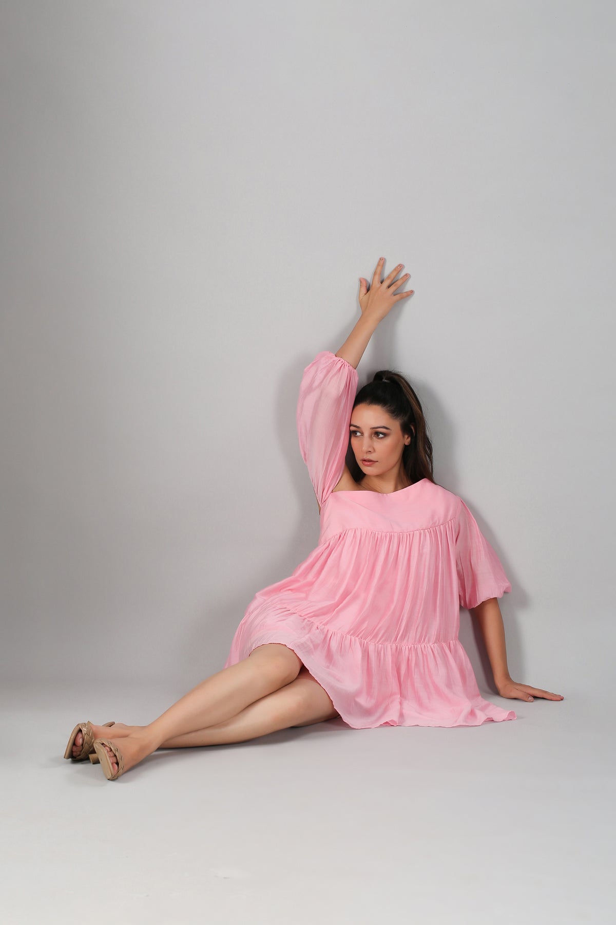 Woman seated against gray backdrop wearing a pink off shoulder dress in flowy tiered mini style and sandals