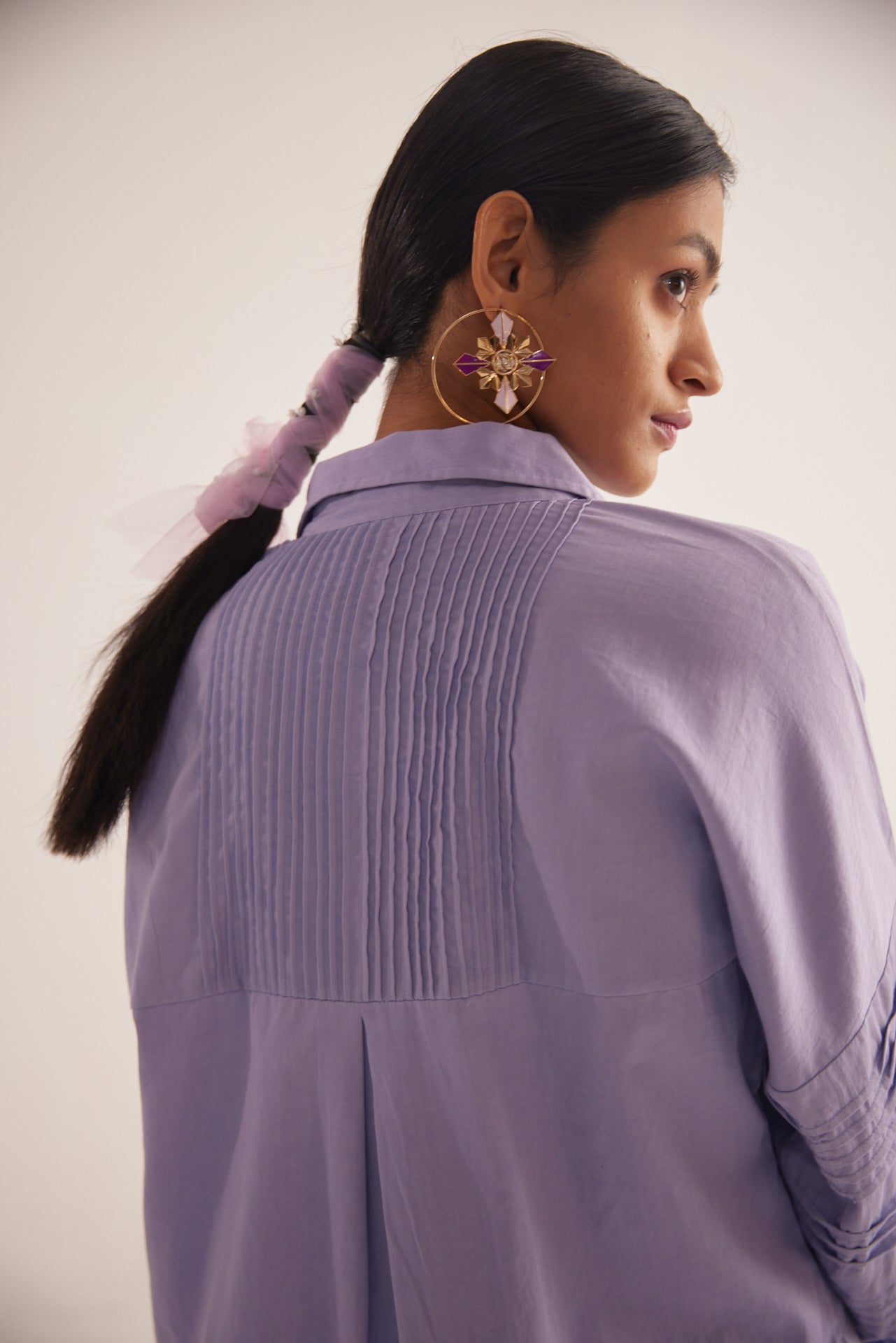 Back view of a woman wearing a purple oversized cotton shirt by Stylera with detailed pleats on the back