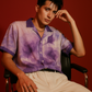 Stylera model wearing purple resort shirt for men floral print short sleeves collar seated in chair against red background