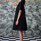 Side profile model wearing a relaxed fit black dress for women with short sleeves textured A line silhouette and black flats