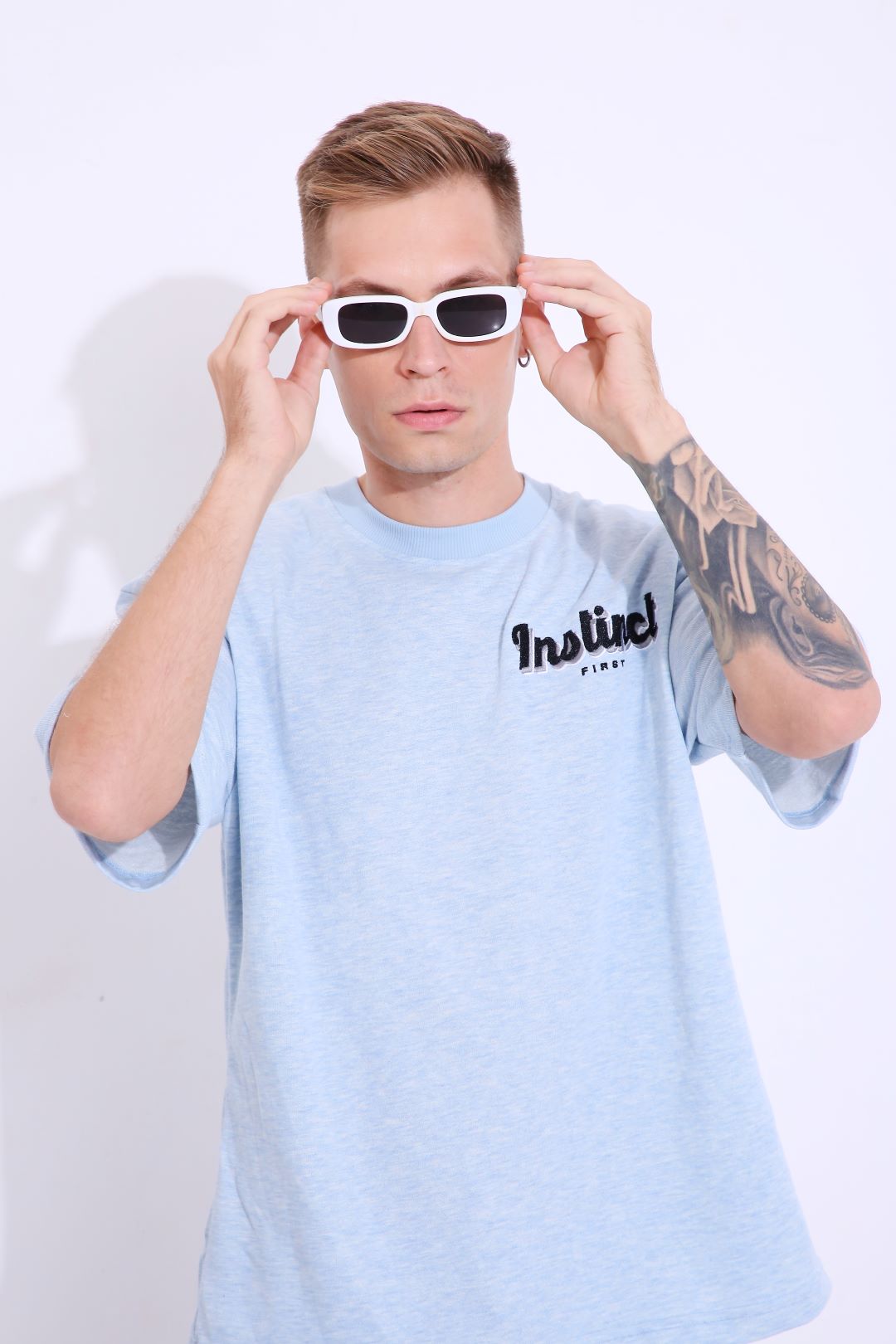 Male model wearing Stylera retro blue oversized t-shirt with logo white sunglasses and visible tattoo