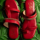 Stylish red flat sandals by Stylera with gold chain detail on green leaves background
