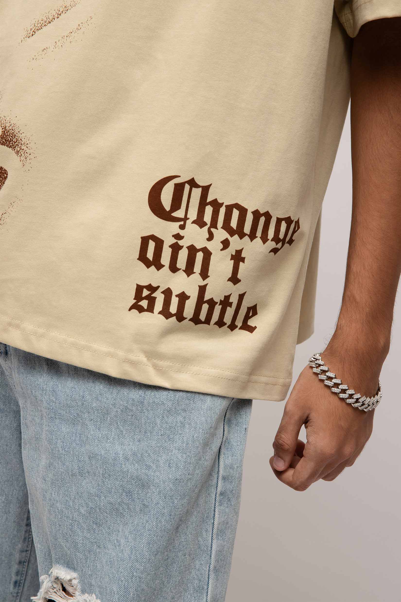 Stylera beige stencil art t-shirt with brown gothic Change aint subtle print distressed hem and model wearing jeans