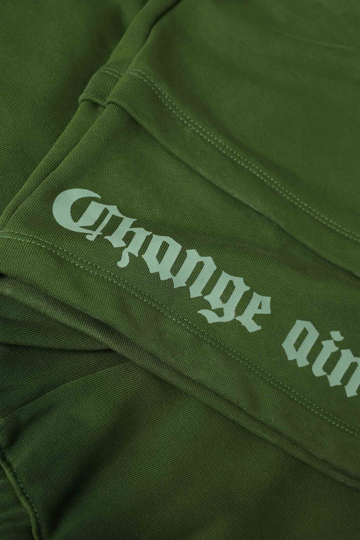 Stylera olive green cargo pocket joggers closeup showing folded fabric seam and white gothic text print on leg