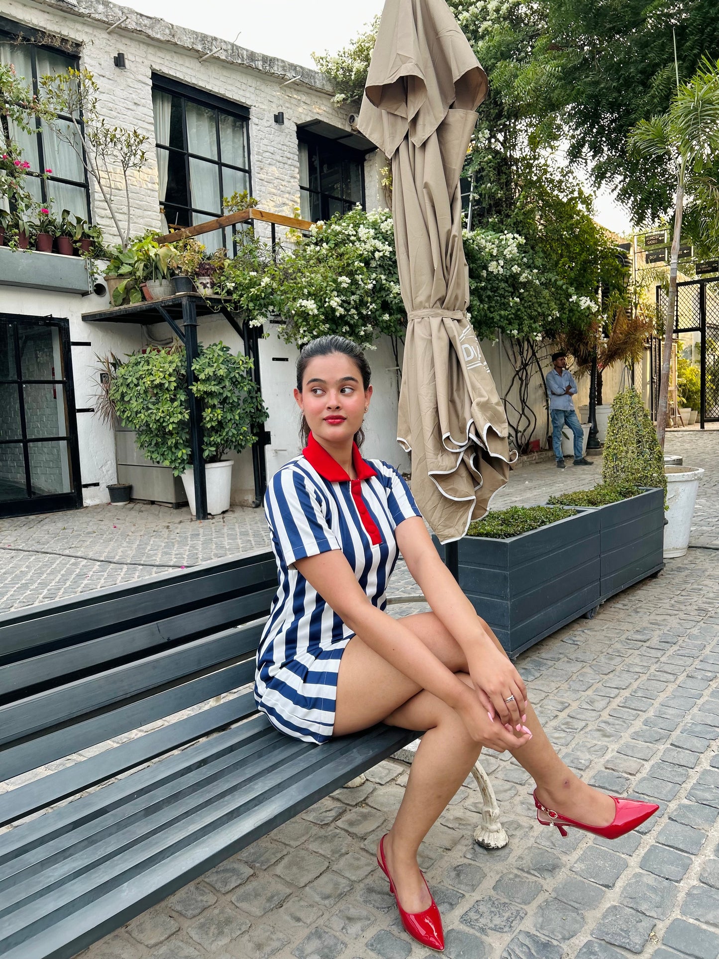 Young woman seated on a bench in a courtyard wearing striped mini dress for women with red collar and red heels
