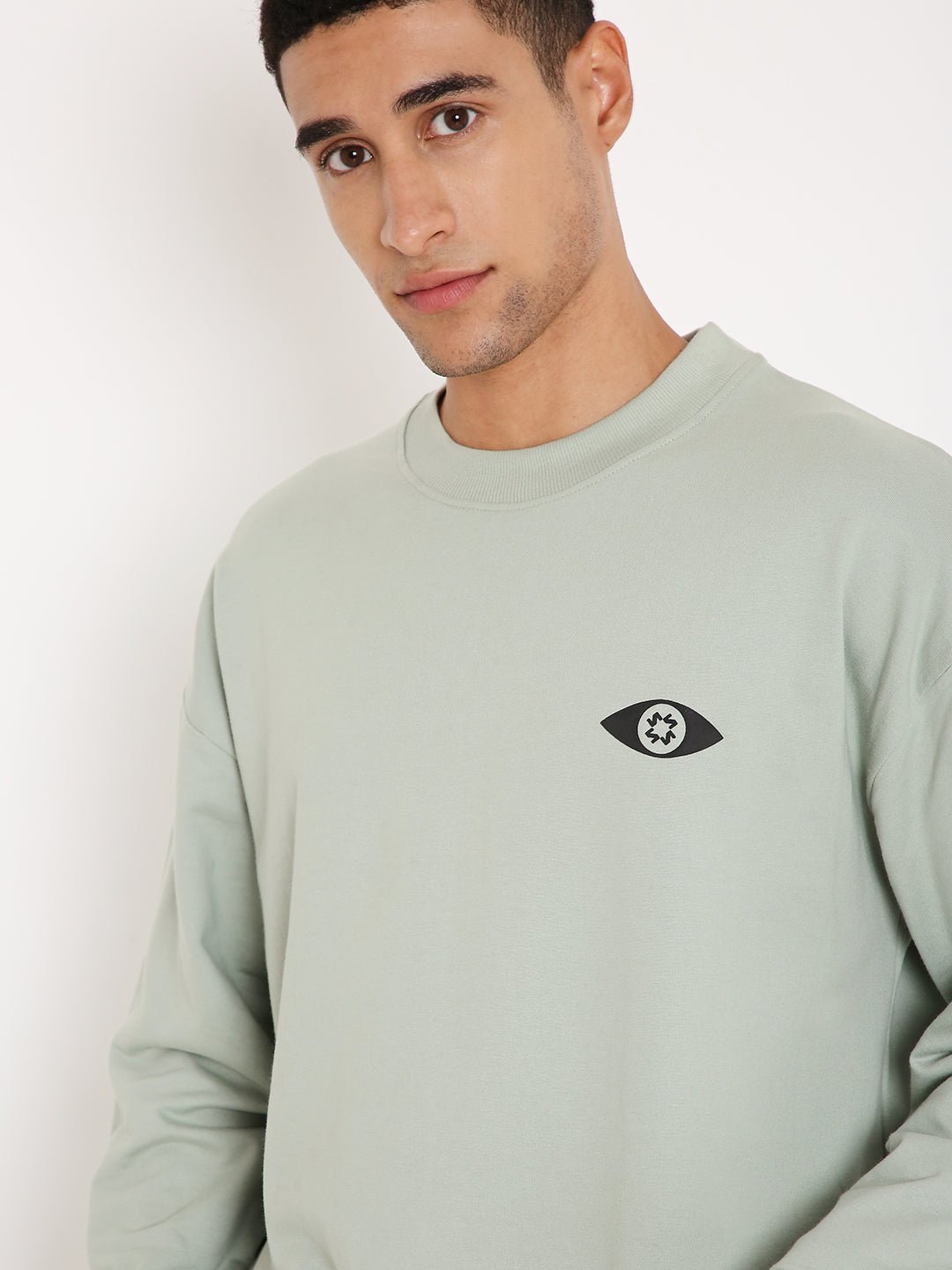 Stylera crewneck sage sweatshirt with small eye logo on chest worn by male model stylish graphic sweatshirts for camping