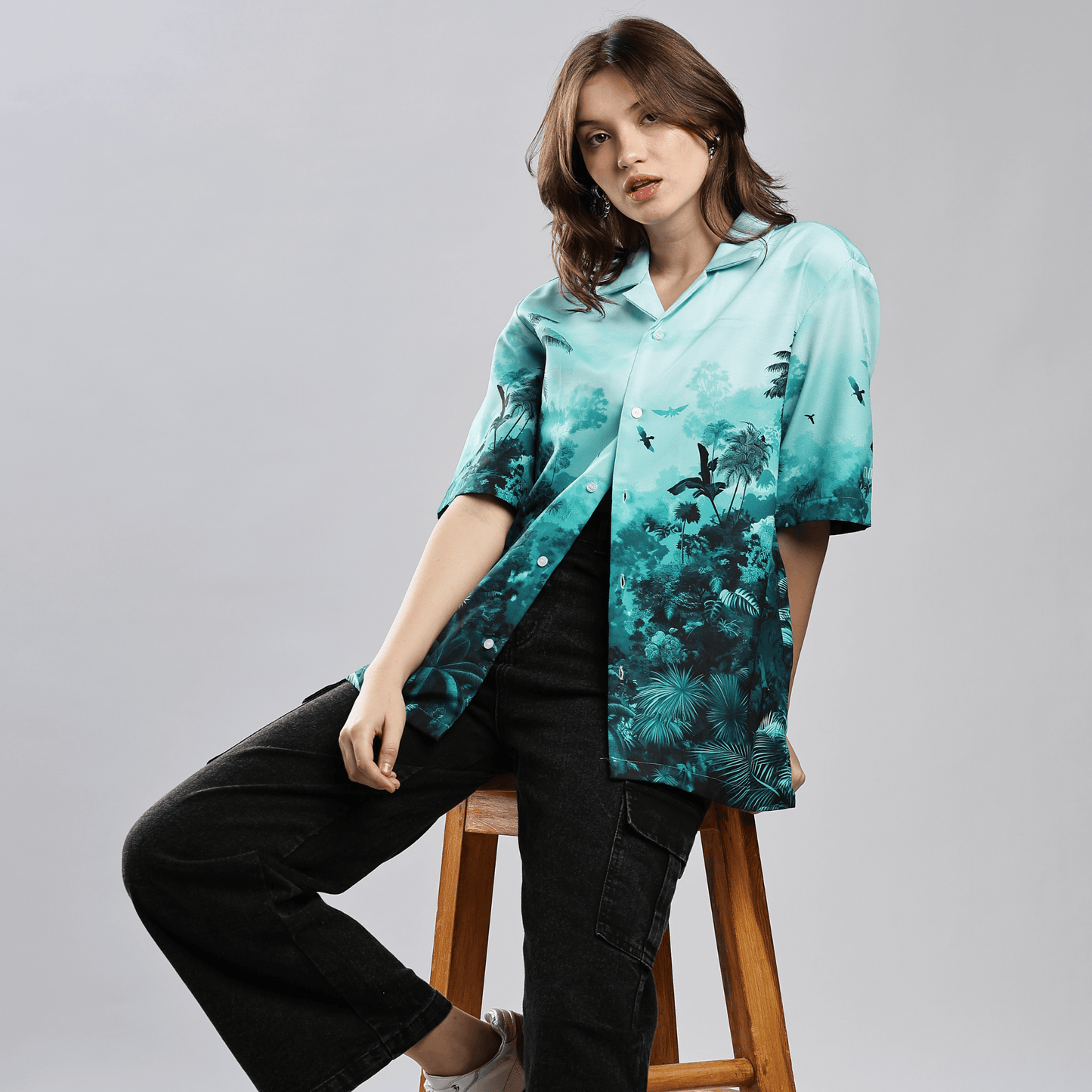 Woman wearing Stylera stylish short sleeve resort shirt turquoise tropical print seated on wooden stool