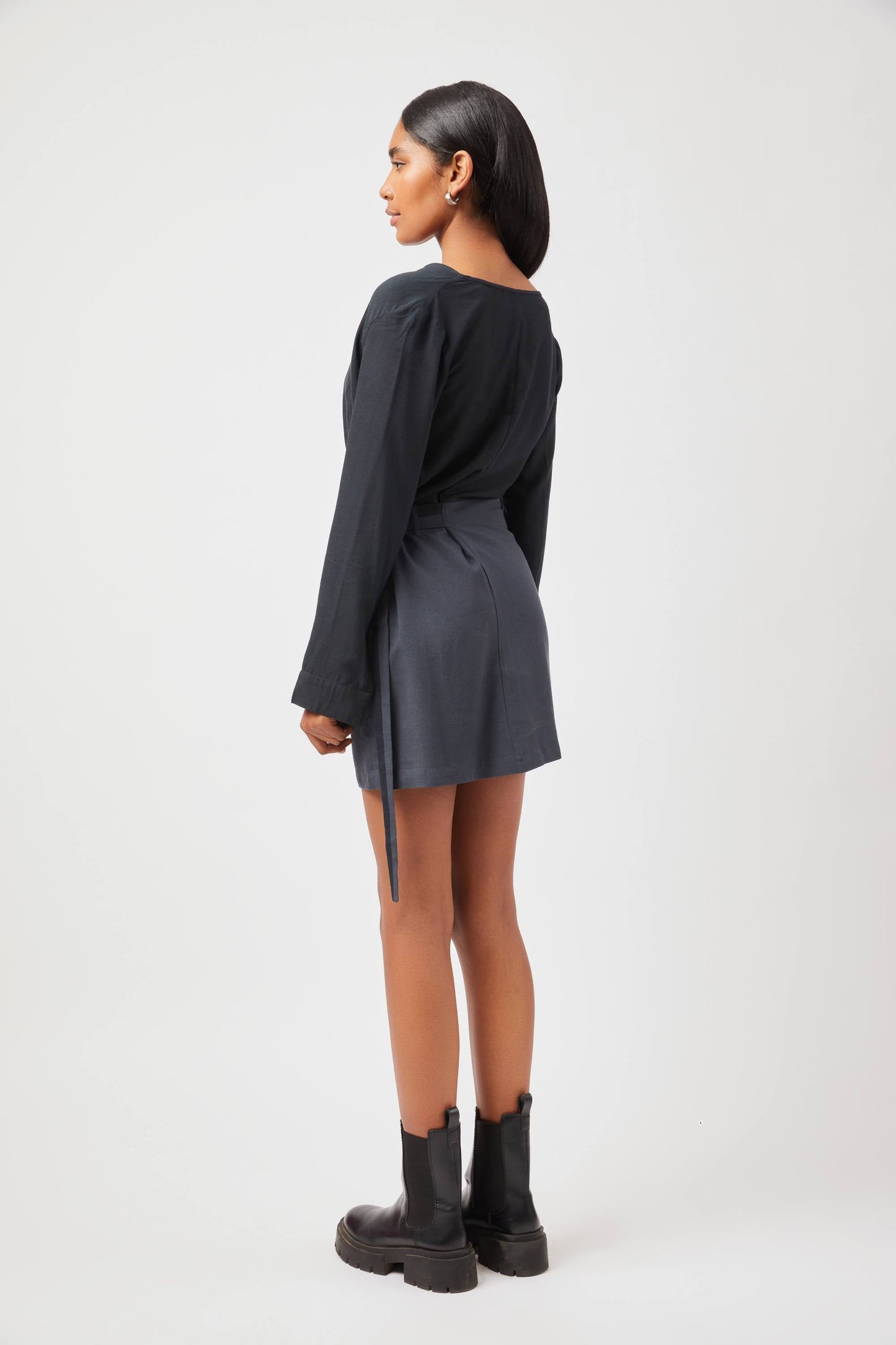 Back view of model wearing Stylera Tate cowl neck dress in black with long sleeves and ankle boots