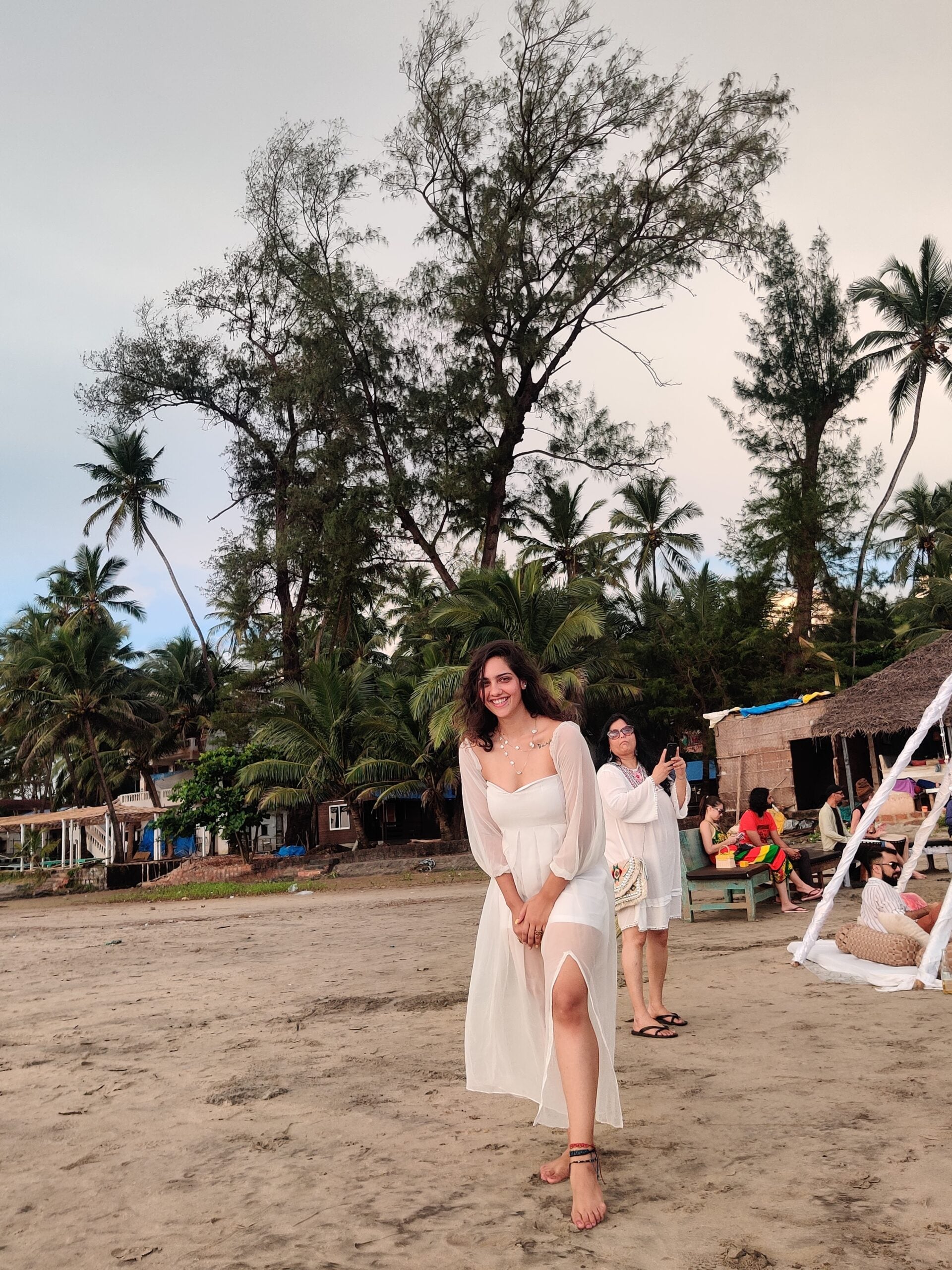 Model wearing a trendy evening maxi dress with a thigh-high slit, walking on a beach surrounded by palm trees