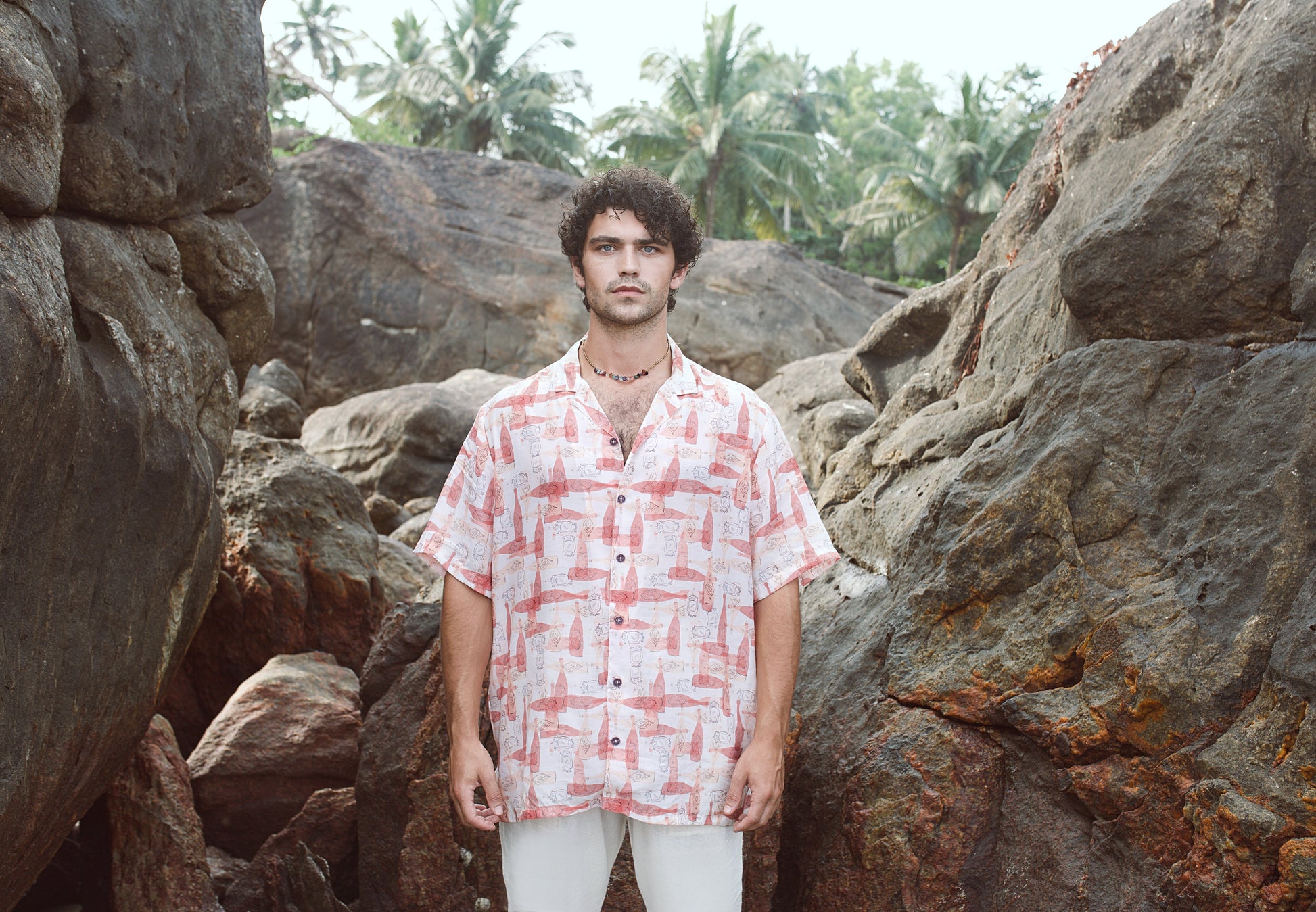 Trendy oversized casual shirt in pink and white worn by a model standing among rocks and palm trees