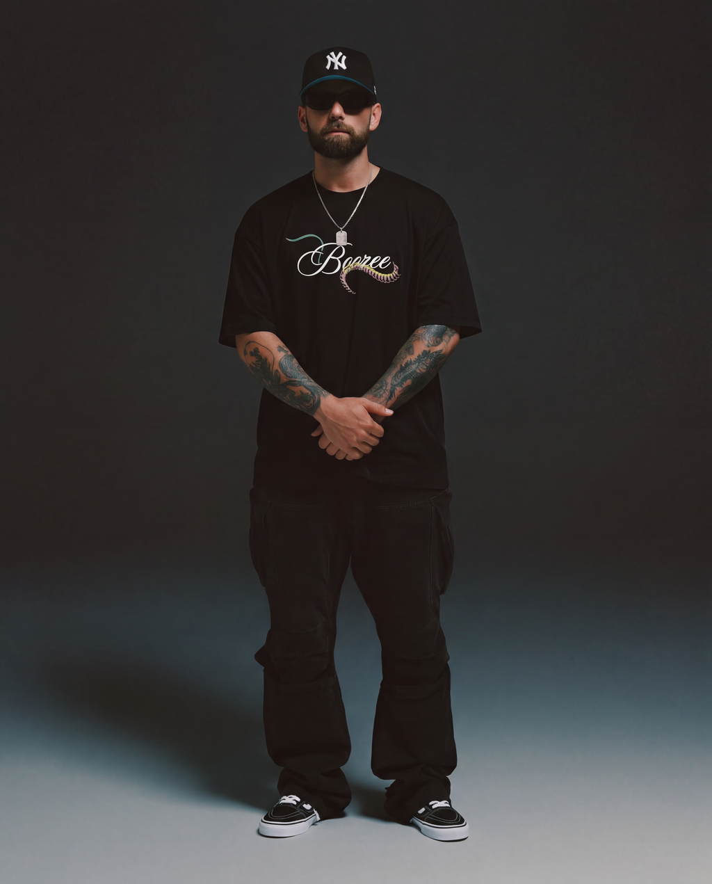 Man wearing a black t-shirt with a logo, black pants, and a black cap on a dark background
