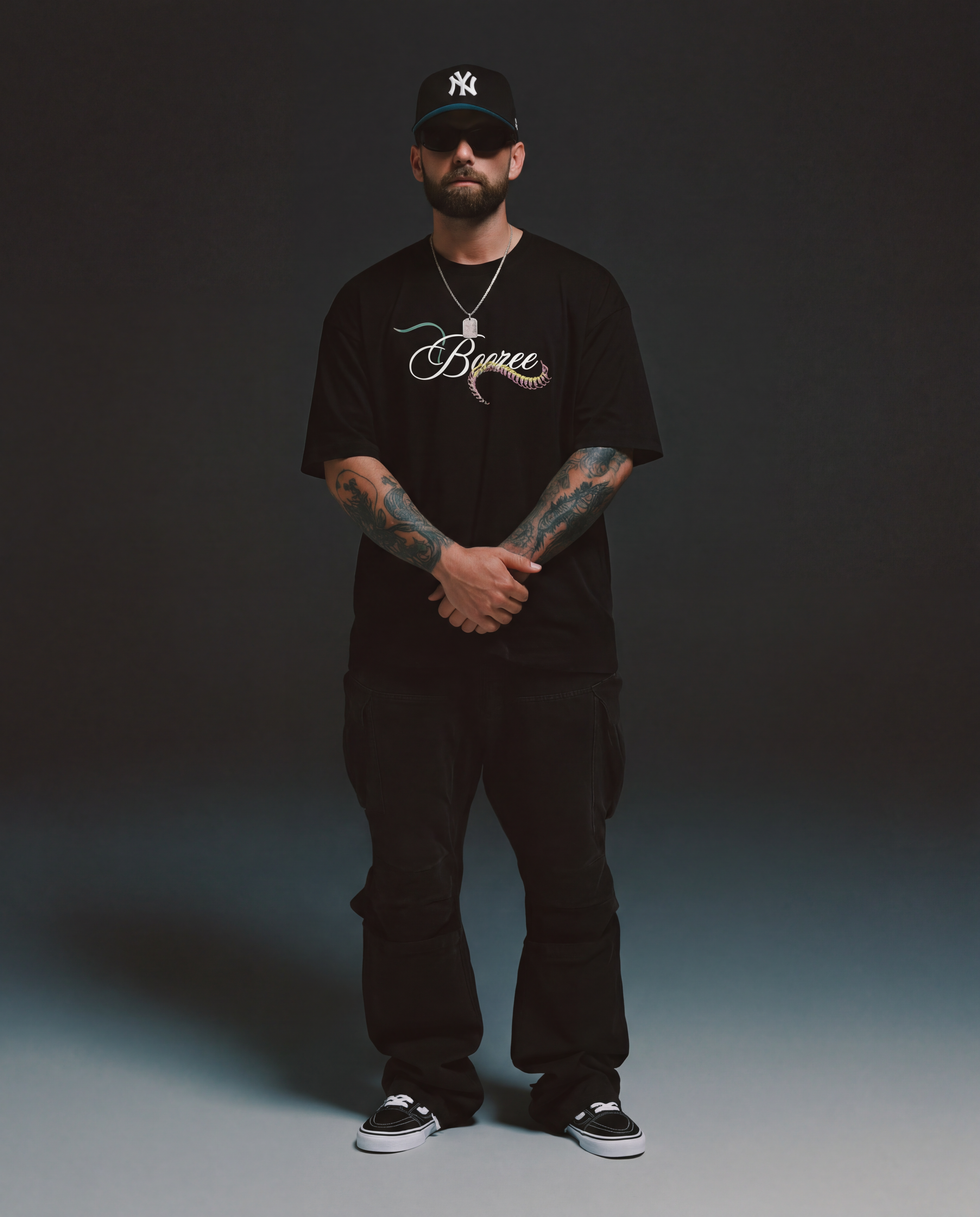 Man wearing a black t-shirt with a logo, black pants, and a black cap on a dark background