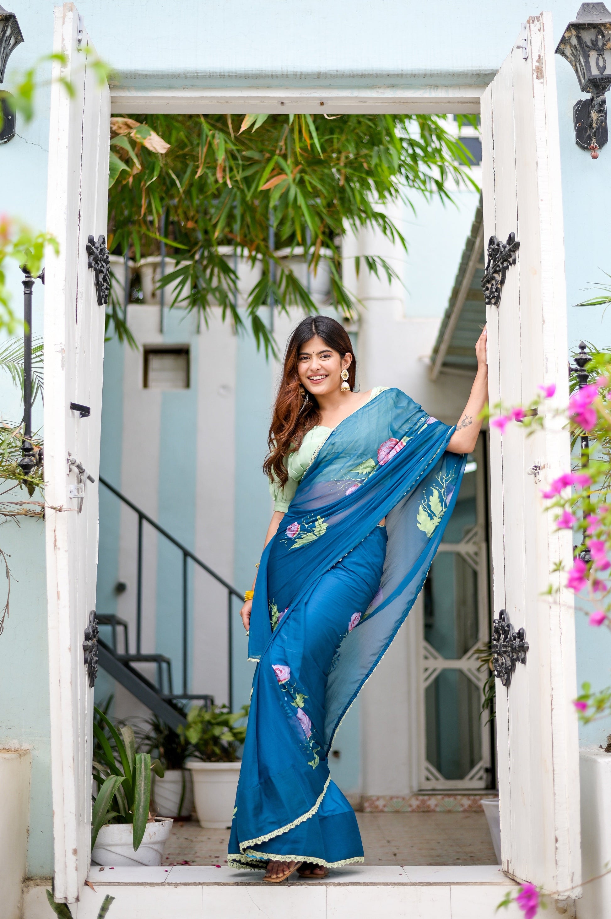 Elegant woman in a unique blue saree for special occasions, adorned with floral patterns, standing in a beautiful entryway
