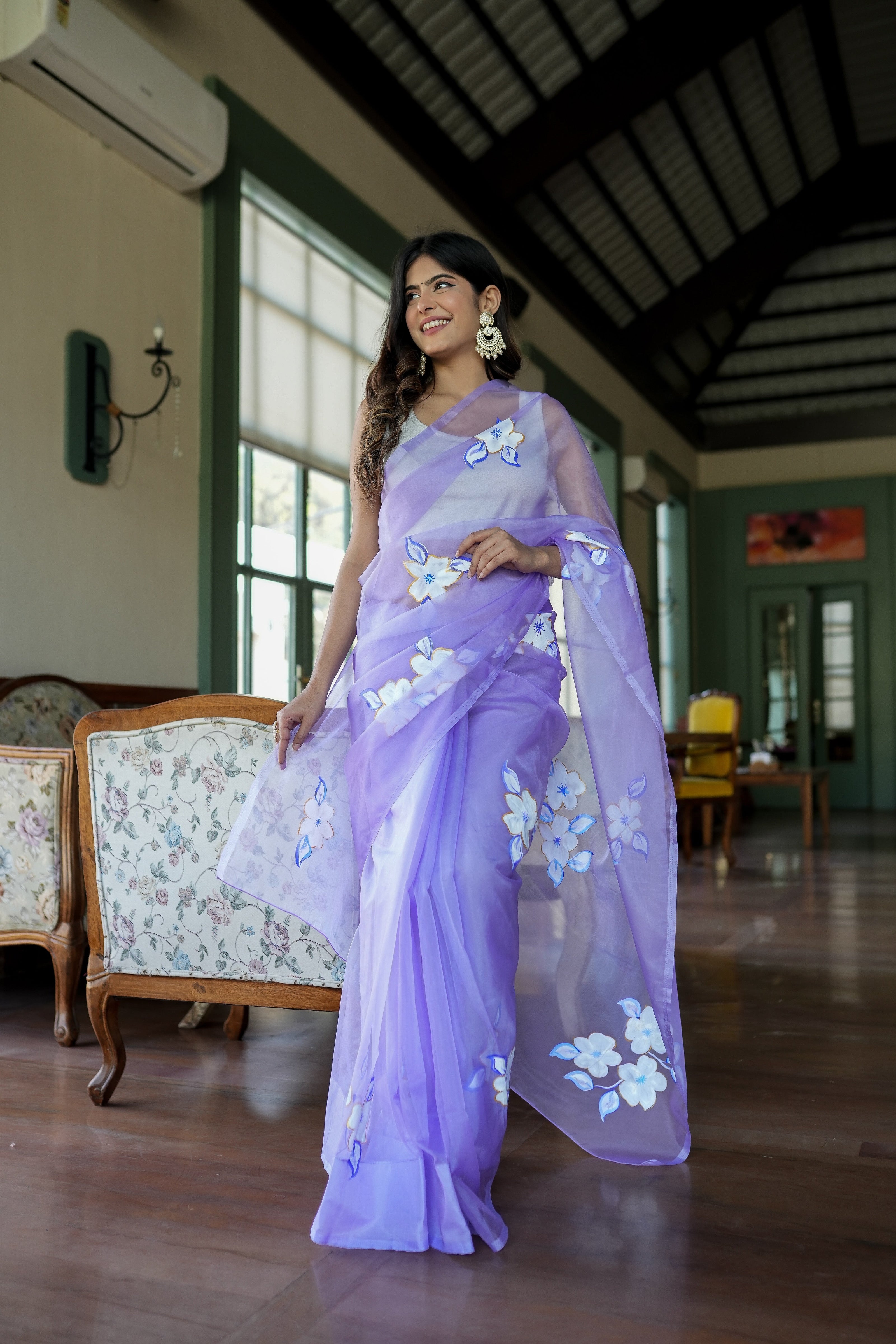 Elegant woman wearing a unique organza saree with artwork in soft lavender, styled for a chic indoor setting.