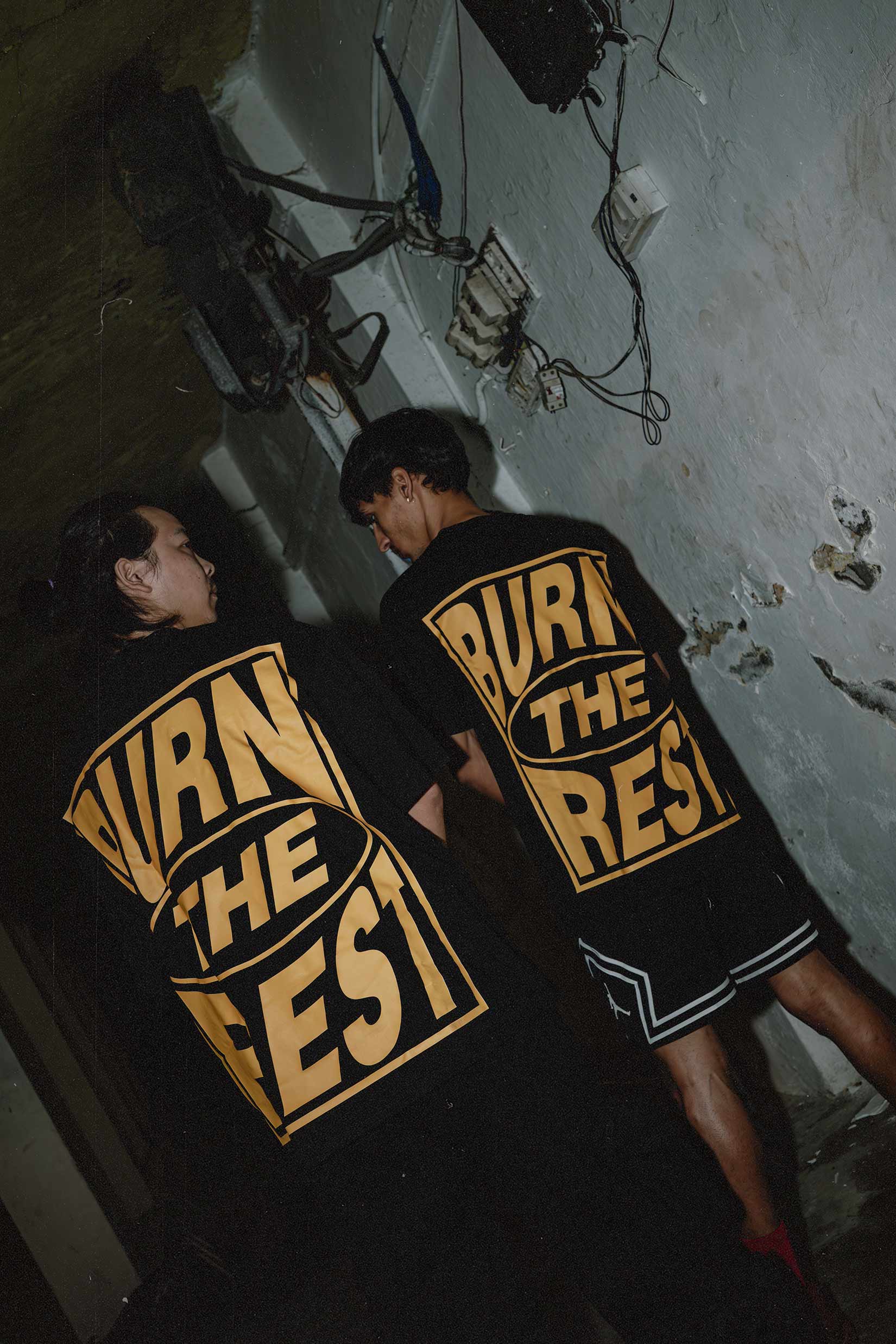 Stylera black graphic t-shirt trendy oversized back print with bold yellow BURN THE REST text worn by two models in gritty hallway