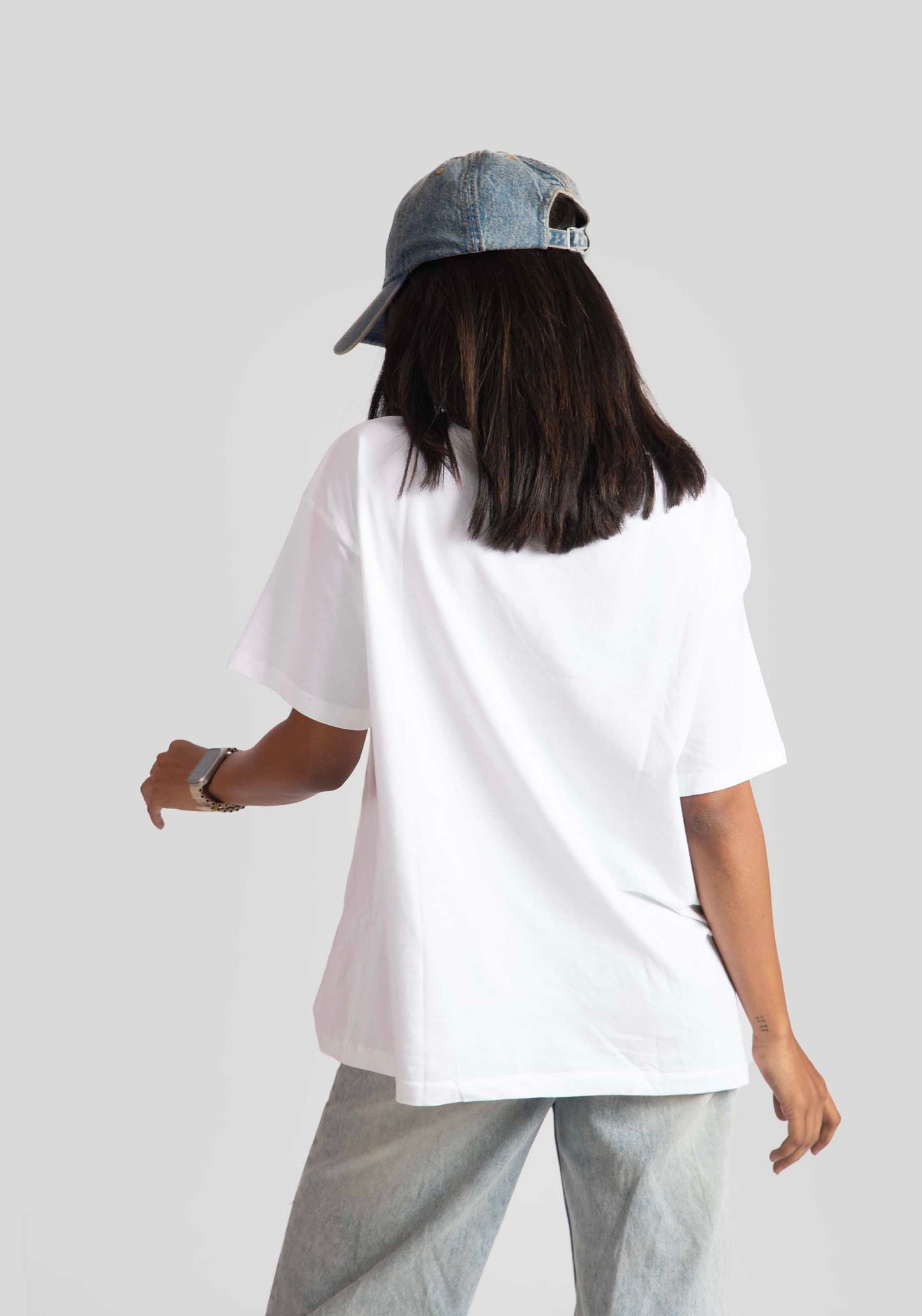 Back view of woman wearing a white good vibes only graphic tee for women with denim cap and light jeans