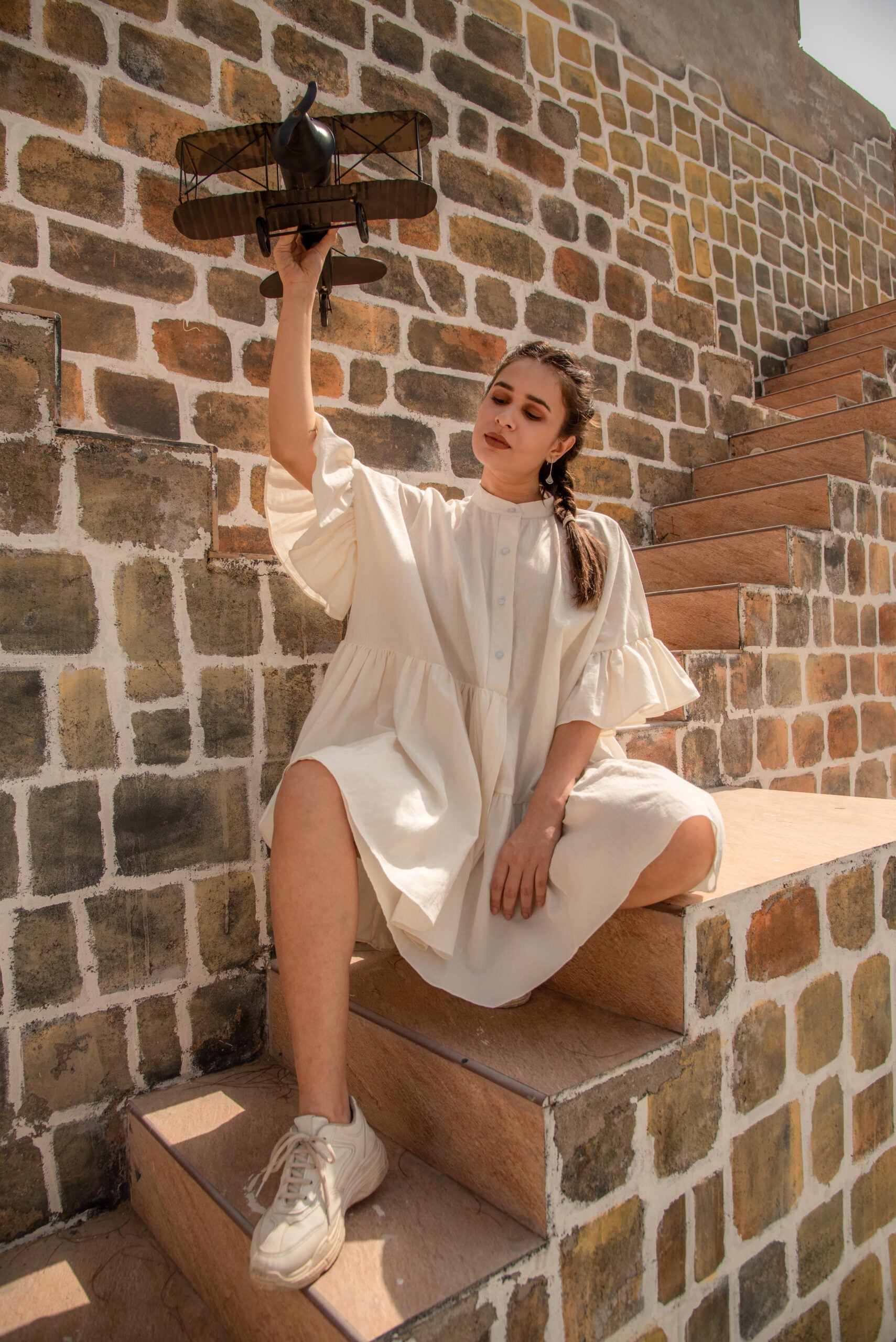 Model wearing a white summer dress for women while holding a vintage airplane on outdoor stairs