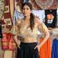 Woman wearing a yellow floral off shoulder crop top with black pants in a colorful market setting