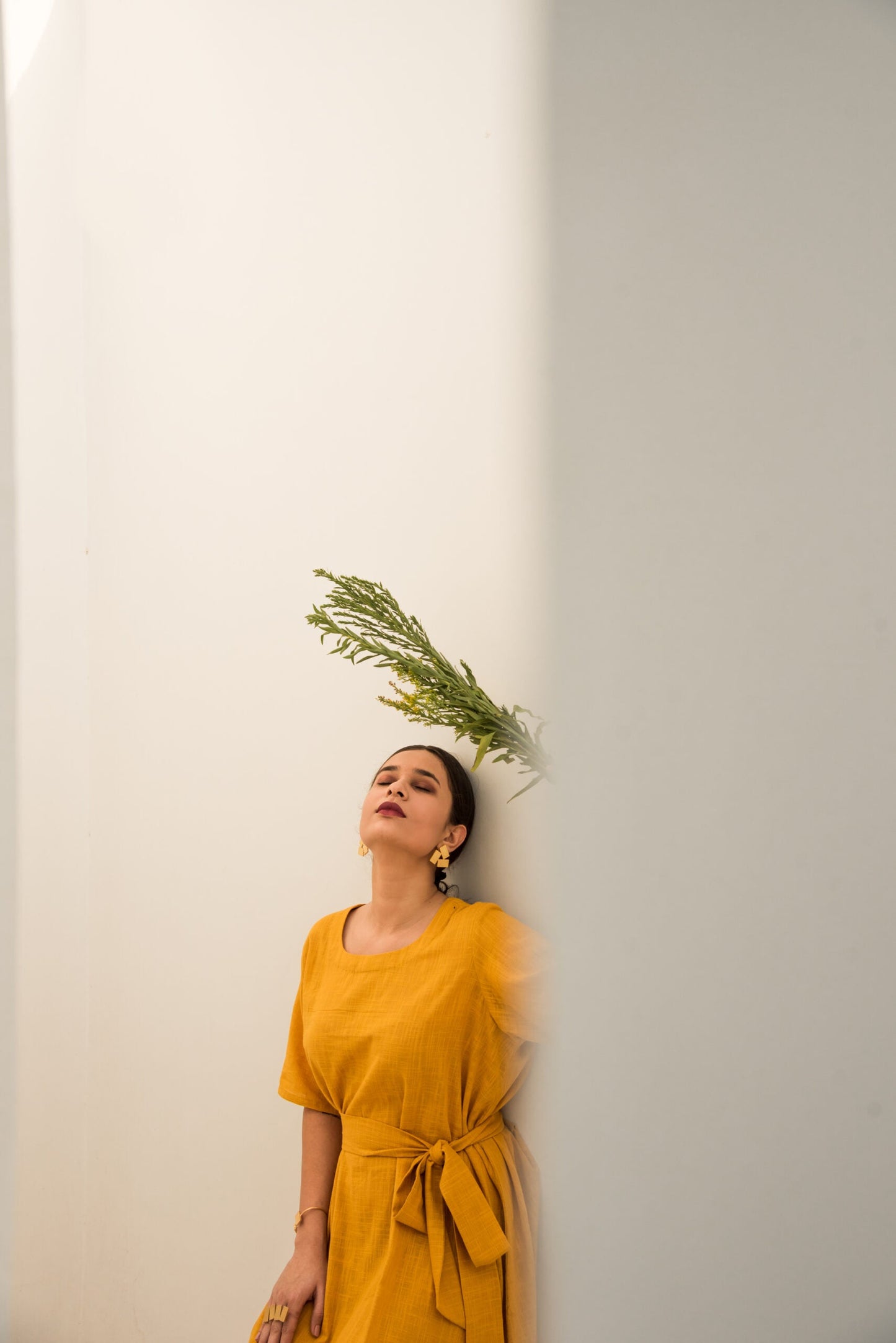 Woman wearing a yellow summer midi dress standing against a white wall with greenery above her head