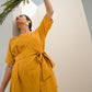 Model wearing a yellow summer midi dress with a tie waist, posing with a plant against a minimalist background