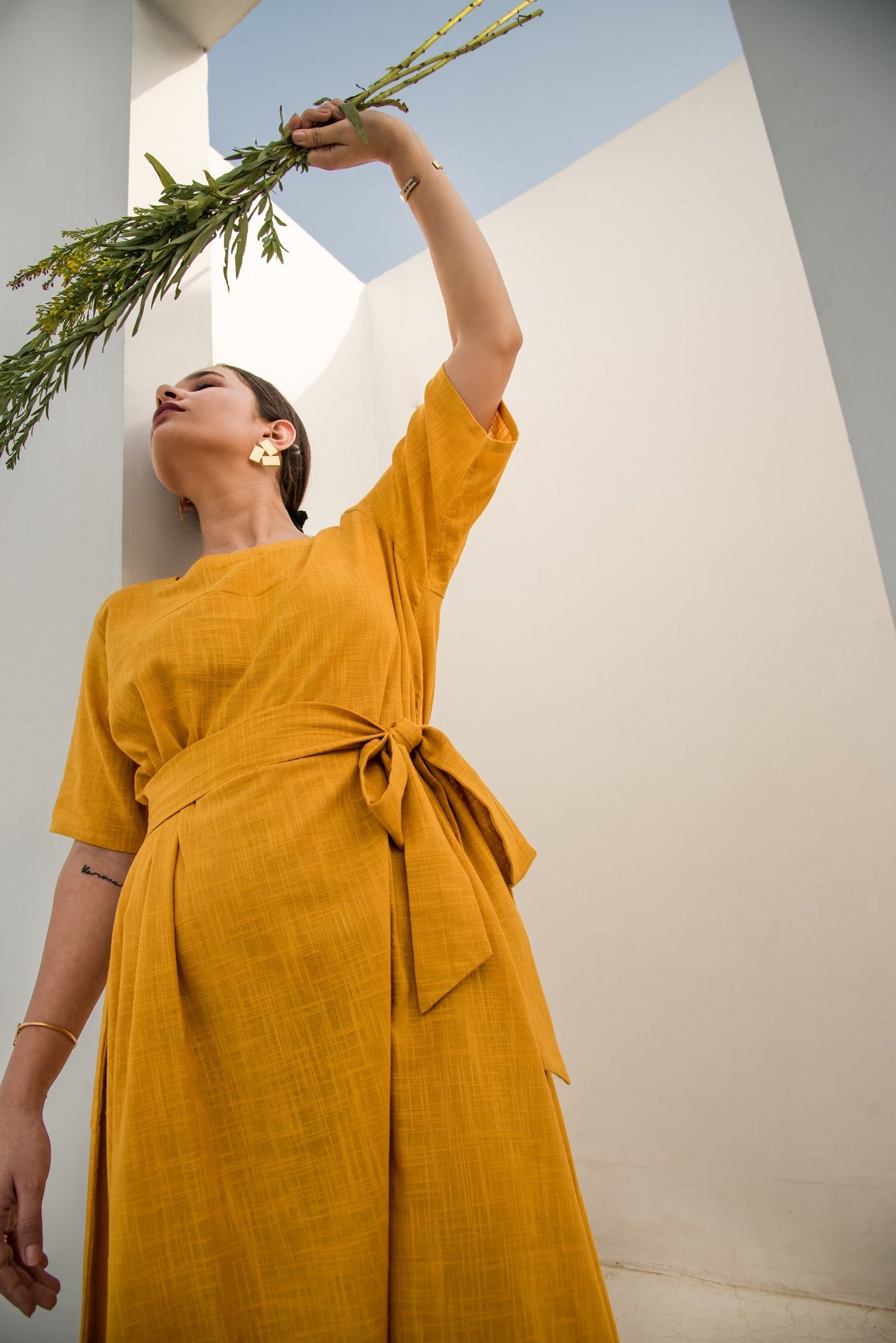 Model wearing a yellow summer midi dress with a tie waist, posing with a plant against a minimalist background